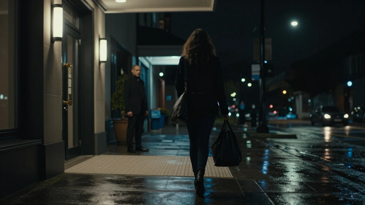 Woman walking away from hotel at night, streetlights glinting, distant figure in background.
